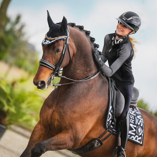 Woman riding a horse wearing. ablack horse riding outifit - black riding tights and a glossy riding helmet. Horse is wearing a black dressage bridle, a fly veil and a black print dressage saddle pad by the brand Mrs Ros