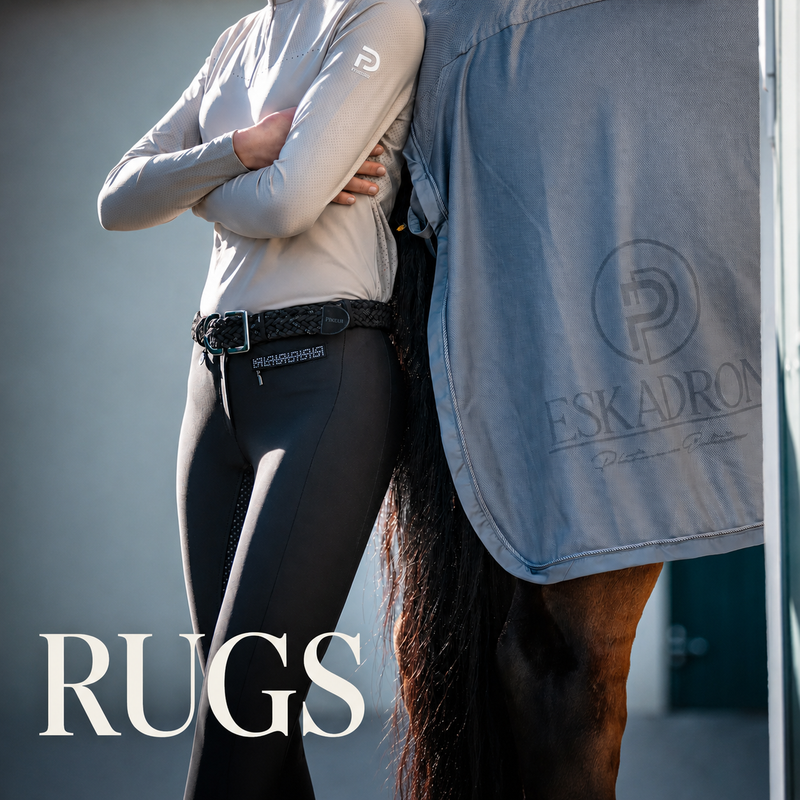 A person in equestrian apparel stands beside a horse wearing a gray ESKADRON rug, with the word 'RUGS' overlaid at the bottom left. The visible apparel includes a light beige long-sleeve shirt, black riding tights, and a black belt.